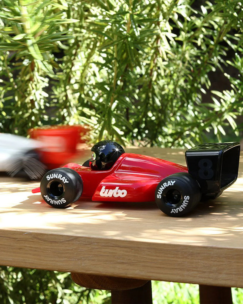 A Playforever Car TURBO Verve Laser Rich Red toy with "turbo" on the side and "SUNRAY" tires sits outdoors on wood, as another blurred racing car toy zooms by in the background amid green foliage.