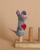 The "Mouse Holding A Heart Finger Puppet" is a cute, handmade in Nepal felt mouse figurine with pink ears, nose, and tail. The delightful mouse stands on a wooden base with two pegs and holds a red heart, set against a beige background.