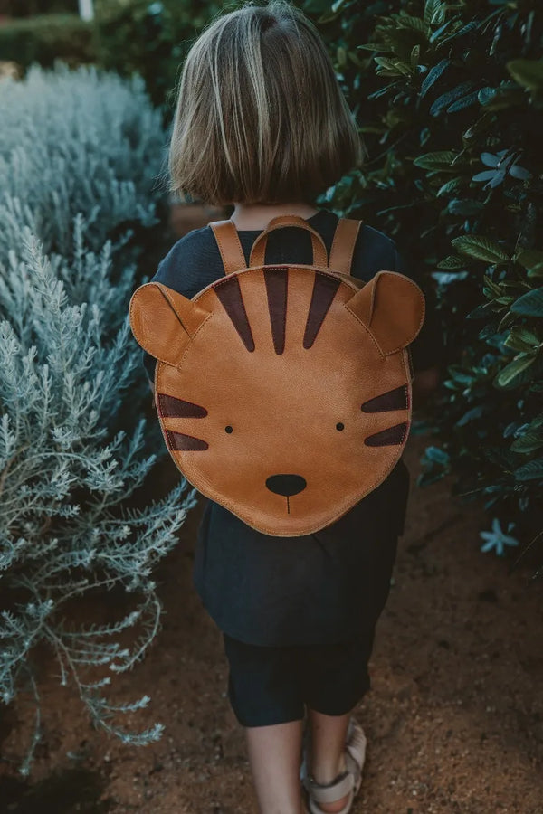 A young child wearing a dark dress and brown sandals, viewed from behind, carries a Donsje School Leather Backpack - Tiger. The child stands between lush green and silver bushes.