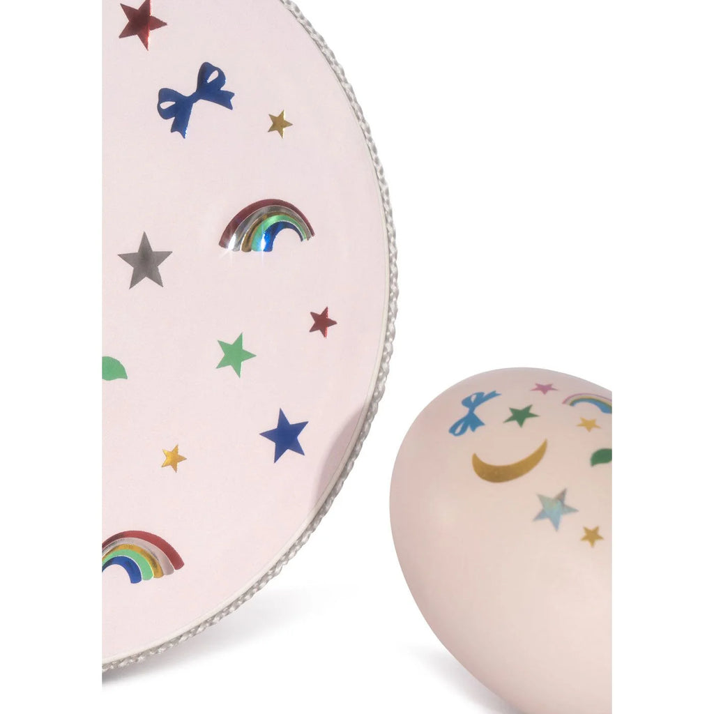 A Konges Sloejd Music Set Petite - Rainbow Twinkle featuring a pale pink circular tambourine and egg shaker, both decorated with colorful metallic rainbows, stars, moons, and bows.