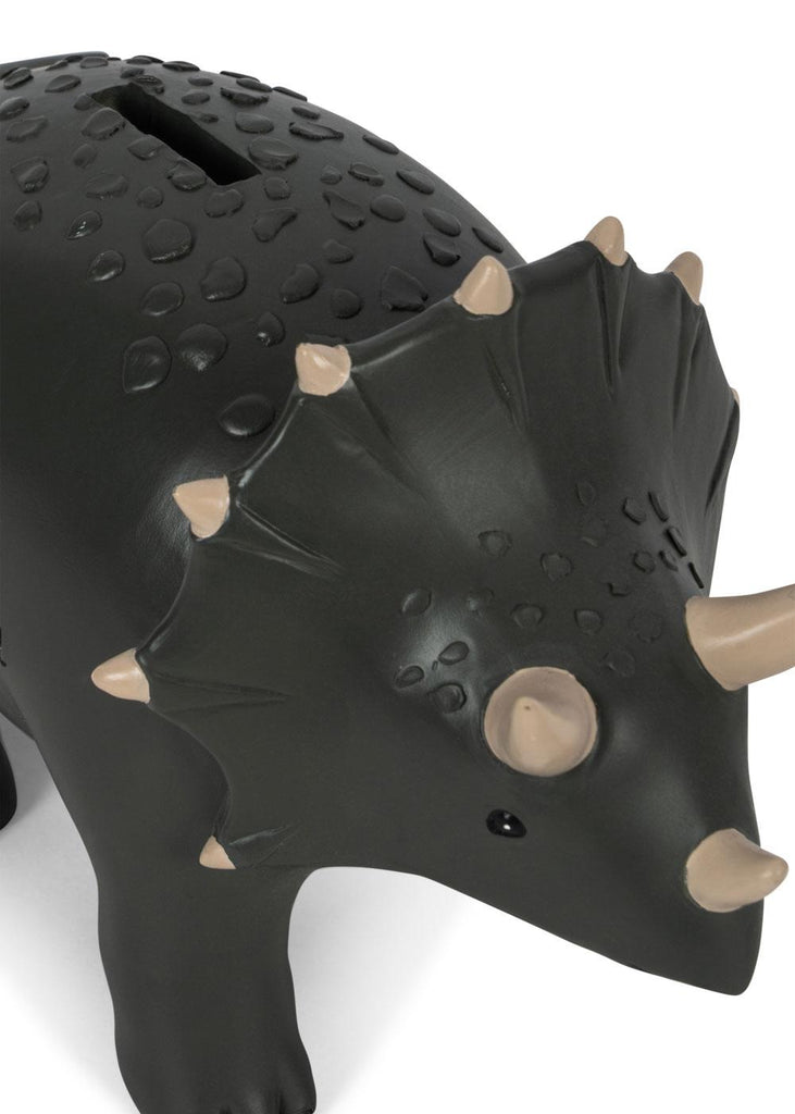 A close-up of the Konges Sloejd Money Bank - Dino, a dark gray triceratops-shaped piggy bank with beige horns and textured skin. The coin slot on its back offers a fun way to store savings.