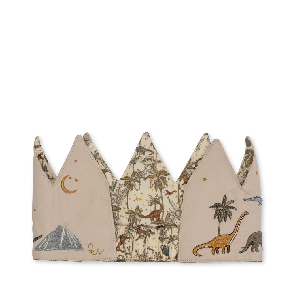 The Konges Sloejd Birthday Crown - Dino features cotton fabric with pointed peaks, embroidered dinosaurs, trees, stars, a crescent moon, and a volcano in earthy tones on a beige background.
