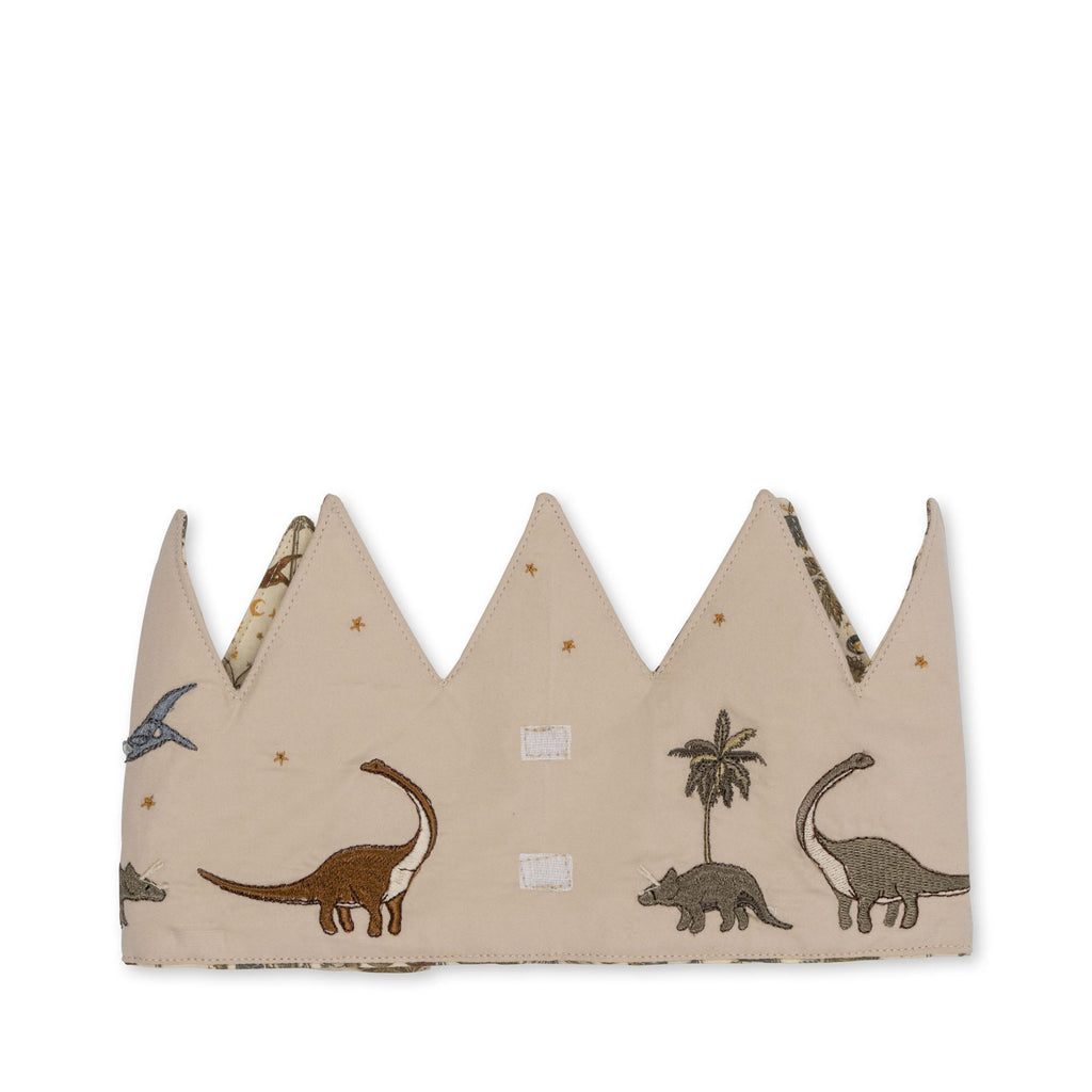 The Konges Sloejd Birthday Crown - Dino is a beige cotton crown with pointed tips, embroidered dinosaurs, palm trees, and stars. It’s adjustable with two central Velcro patches for easy fastening.