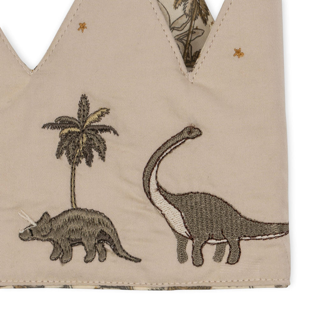 The Konges Sloejd Birthday Crown - Dino is a beige cotton crown with embroidered palm tree, triceratops, and long-necked dinosaur. It features a zigzag shape, gold stars, and an adjustable fit—perfect for special birthday celebrations.
