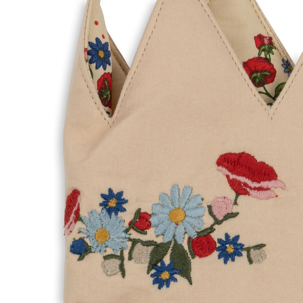 A close-up of the Konges Sloejd Birthday Crown - Flower, a beige fabric crown adorned with colorful embroidered blue daisies, red poppies, white flowers, and green leaves along the base and tips.