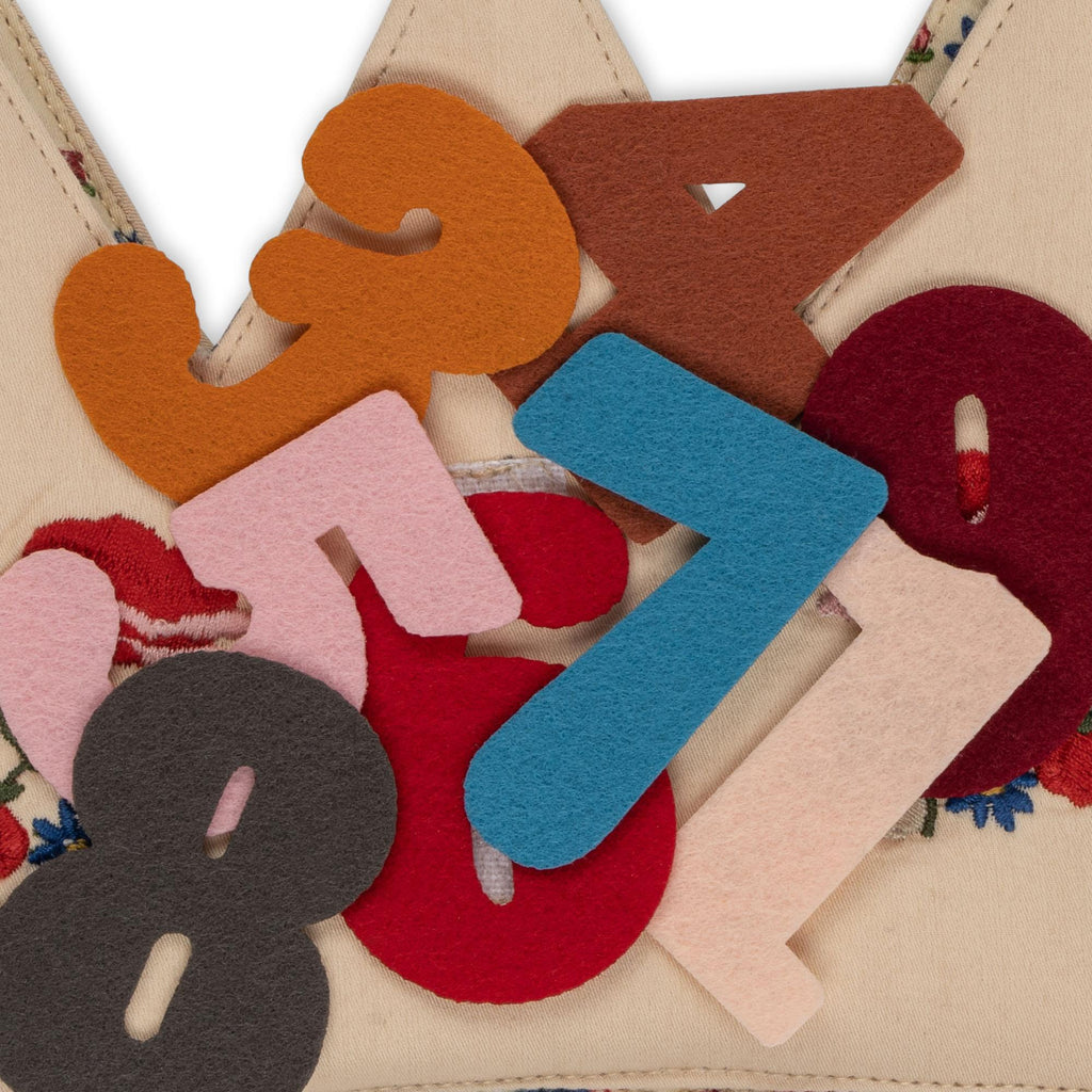 A pile of colorful felt numbers (3-8) overlaps on a beige, floral-embroidered fabric—the Konges Sloejd Birthday Crown - Flower, perfect for decorating a birthday crown or hat.