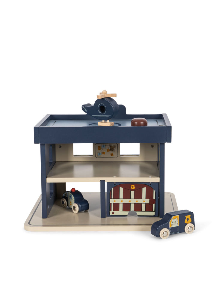 The Konges Sloejd Wooden Police Station is made from FSC-certified wood and features a blue and beige garage, a rooftop toy helicopter, two police cars, a jail door, and an illustrated sign.