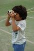 A young child with dark hair, in a white t-shirt and blue shorts, stands on an outdoor sports court looking through Konges Sloejd Explorer Binoculars - Lemon.