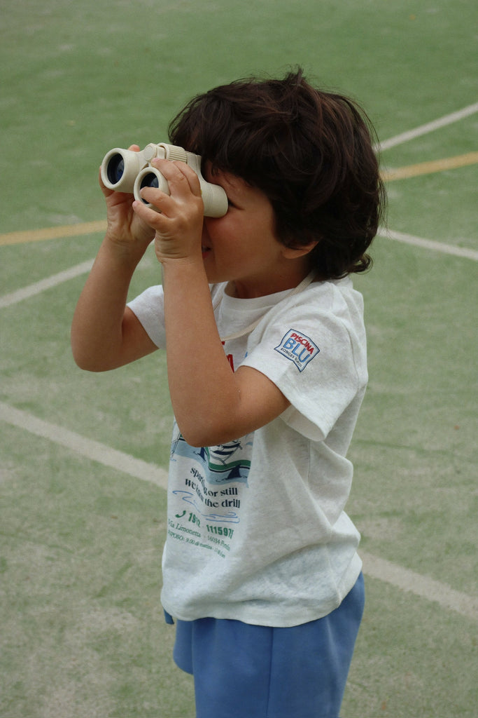 A young child with dark hair, in a white t-shirt and blue shorts, stands on an outdoor sports court looking through Konges Sloejd Explorer Binoculars - Lemon.