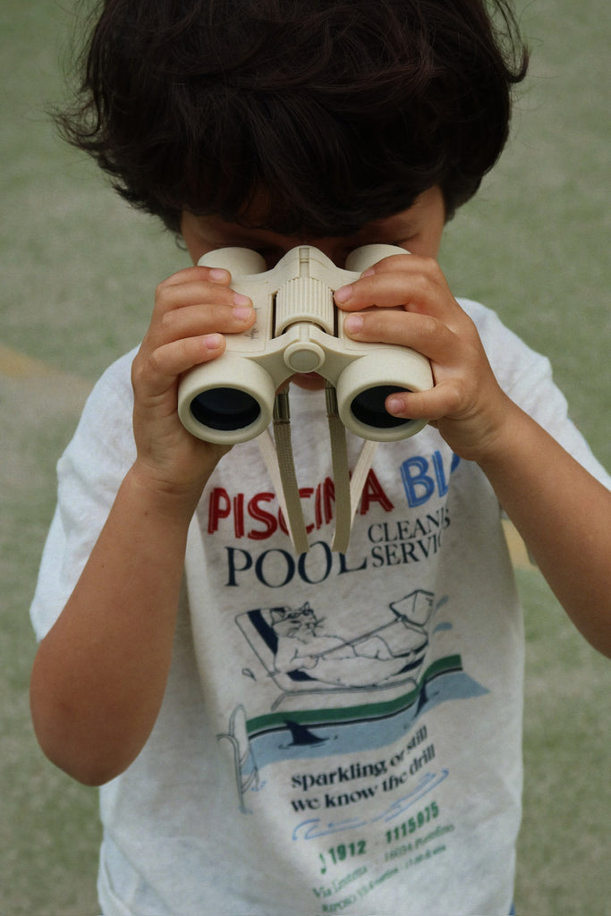 A dark-haired child uses Konges Sloejd Explorer Binoculars - Lemon, peering through them. They're wearing a white t-shirt with colorful graphics, with a blurred outdoor background behind.