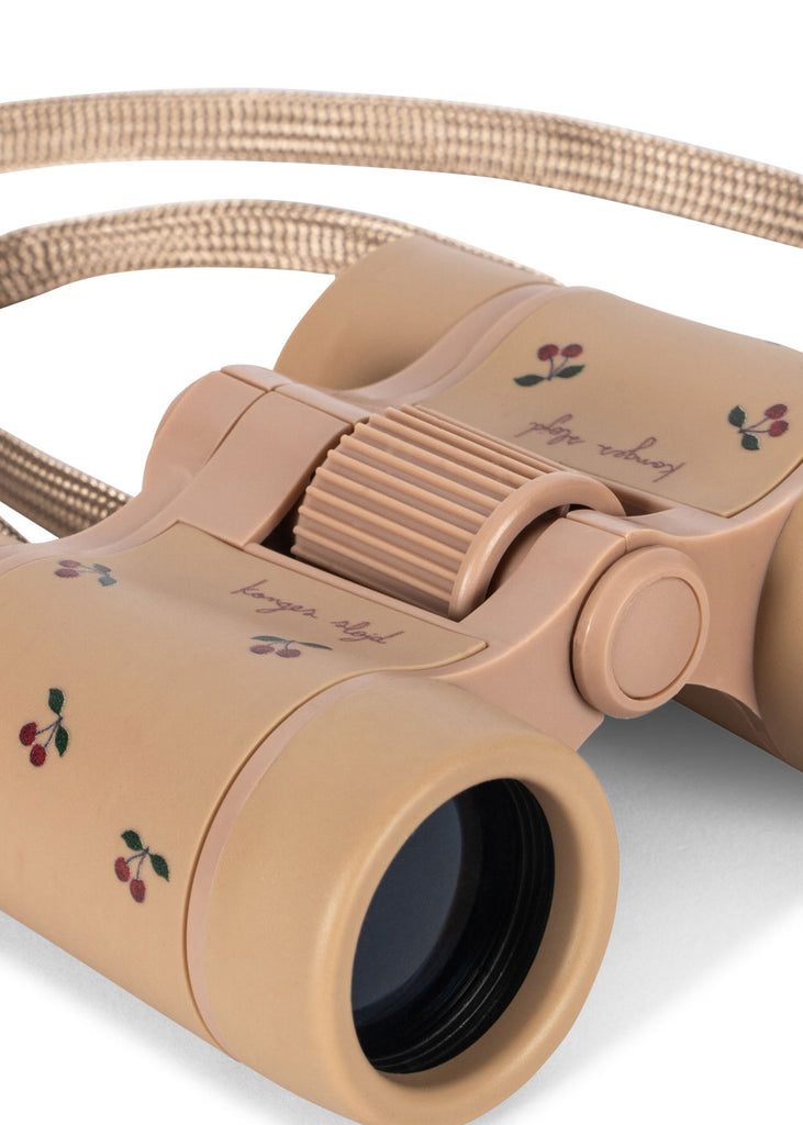 A close-up of Konges Sloejd Explorer Binoculars - Cherry, featuring a lightweight beige design with adjustable focus, a braided strap, small cherry illustrations, and cursive writing.