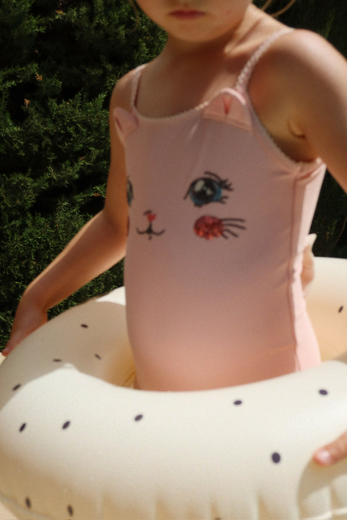 A child in a pink swimsuit with an animal face holds the Konges Sloejd Junior Swim Ring - Navy Dot while standing outdoors in sunlight. The child’s face is partially out of frame.