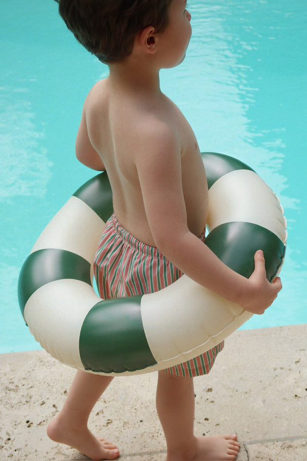 A young boy in striped swim trunks stands by a pool, wearing the Konges Sloejd Junior Swim Ring in Green Stripe around his waist.