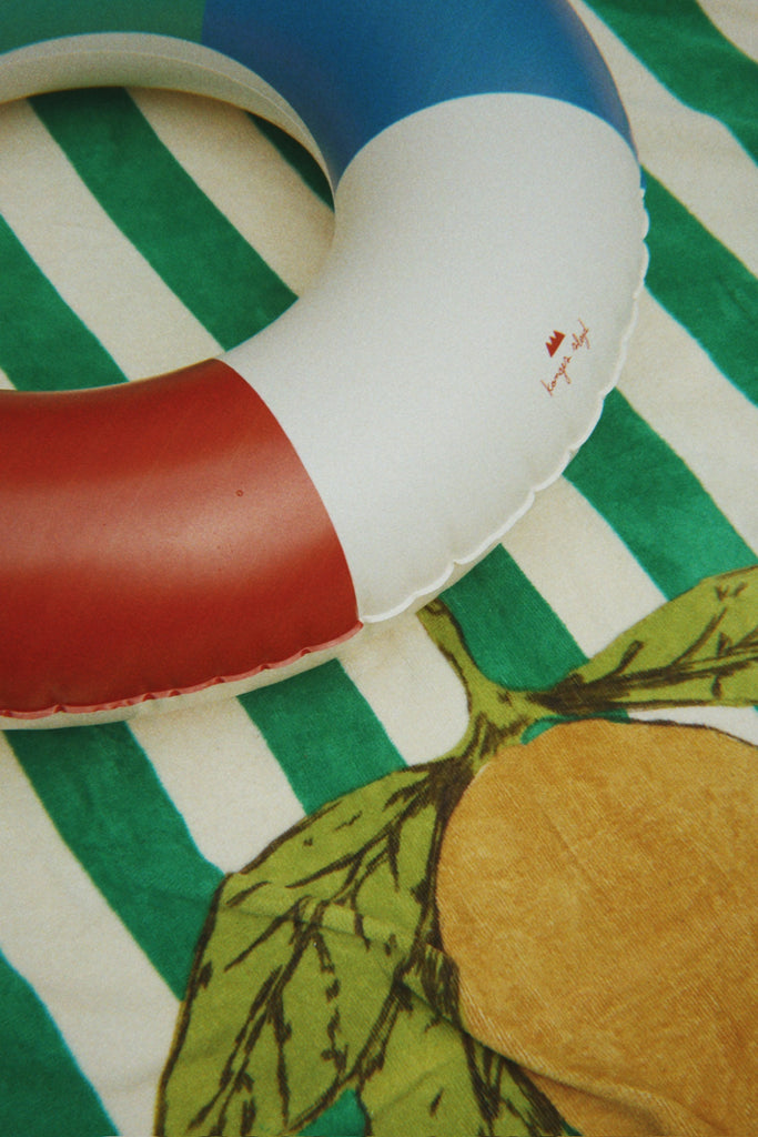 The Konges Sloejd Junior Swim Ring - Multi adds a playful touch to summer fun, shown resting on a green and white towel decorated with a large yellow lemon and green leaves.