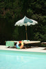 A poolside scene with two wooden lounge chairs, one draped with a green-and-white striped towel, the Konges Sloejd Junior Swim Ring - Multi as a playful accessory, and a white sun umbrella. Green trees complete the natural backdrop.