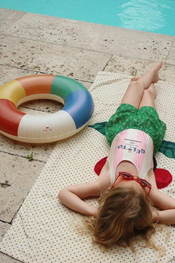 A young child with long hair relaxes on a towel by the pool, wearing a swimsuit and green shorts, with the Konges Sloejd Junior Swim Ring - Multi beside her—a fun and colorful pool accessory—enjoying a sunny day.