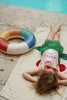 A young child with long hair relaxes on a towel by the pool, wearing a swimsuit and green shorts, with the Konges Sloejd Junior Swim Ring - Multi beside her—a fun and colorful pool accessory—enjoying a sunny day.