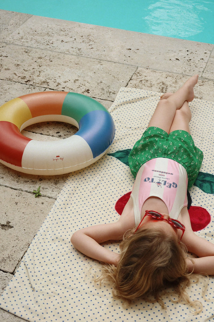 A young child with long hair relaxes on a towel by the pool, wearing a swimsuit and green shorts, with the Konges Sloejd Junior Swim Ring - Multi beside her—a fun and colorful pool accessory—enjoying a sunny day.