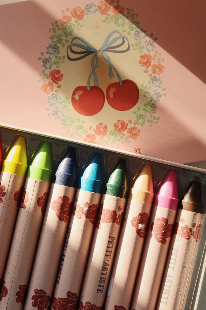 The Konges Sloejd Beeswax Crayons - Cherry Bow come in a floral-decorated pastel pink box, featuring a cherry illustration and blue ribbon on the lid. Sunlight softly highlights the colorful, non-toxic crayons inside.