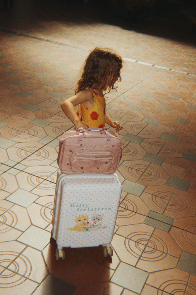 A young child with curly hair in a yellow dress stands on a tiled floor, holding the handle of a white suitcase with cartoon designs and the Konges Sloejd Holiday Essentials Bag - Cherry stacked on top.