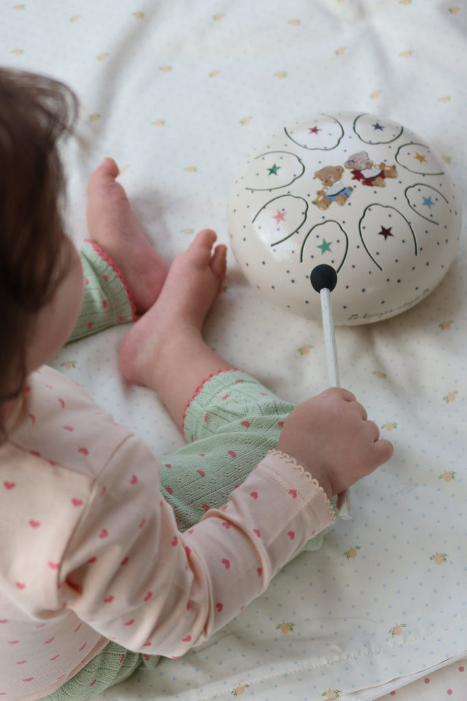 A young child sits on a light-colored blanket, joyfully tapping the Konges Sloejd Marly Steel Drum—a white steel tongue drum—with a black mallet. Dressed in pale pajamas patterned with pink hearts and green pants featuring pink trim, the child offers an ideal example of how beginners can delight in discovering music.