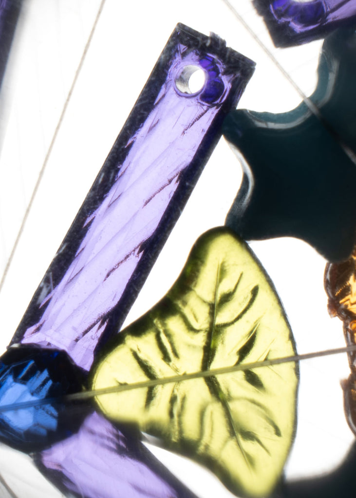 A close-up of the Konges Sloejd Kaleidoscope - Like Cats and Dogs reveals vibrant stained glass-style pieces—including a purple rectangle with a hole, a yellow-green leaf shape, and a teal fragment—all illuminated from behind to showcase their intricate textures and dazzling colors in a mesmerizing kaleidoscopic display.