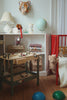 A cozy kids’ room with a red crib, stuffed animals, a Konges Sloejd Tool Bench, a shelf holding books, a lamp, a globe, and a tiger face wall decoration. Two textured balls are on the wooden floor.
