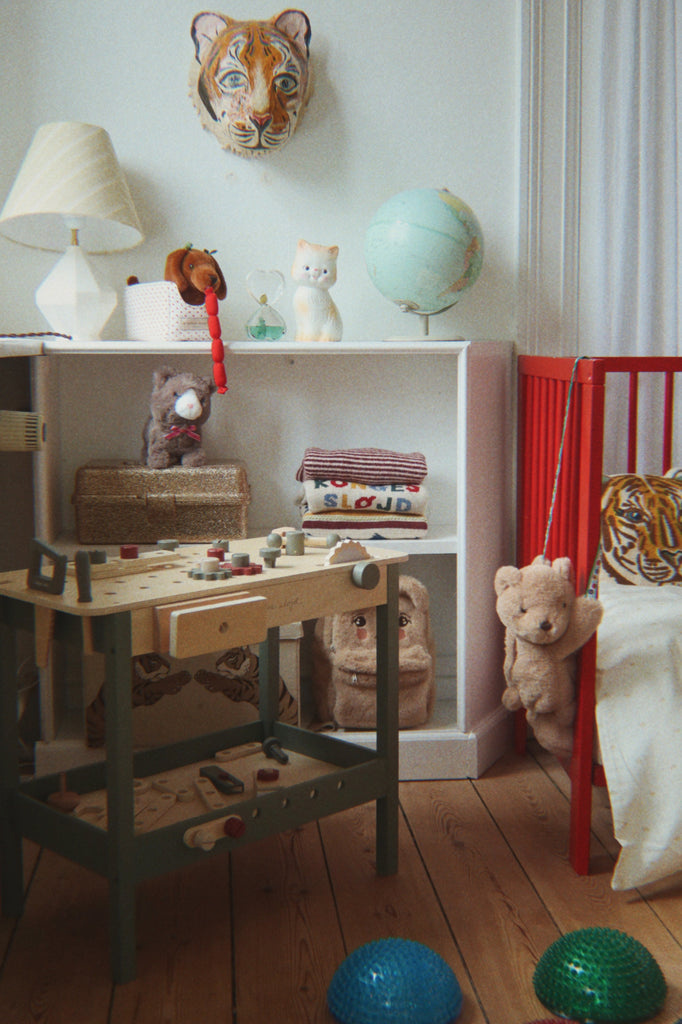 A cozy kids’ room with a red crib, stuffed animals, a Konges Sloejd Tool Bench, a shelf holding books, a lamp, a globe, and a tiger face wall decoration. Two textured balls are on the wooden floor.