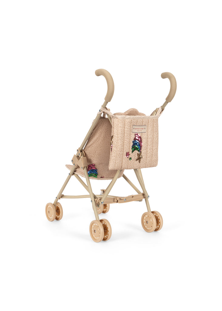 The Konges Sloejd Tulle Doll Stroller - Rainbow Star is a beige stroller featuring curved handles, four pairs of double wheels, and a sparkly rainbow and star patch on the fabric seat. The stroller is displayed at an angle to show both the back and side.