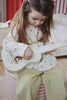 A young girl with long hair sits on a striped pouf and plays the Konges Sloejd Wooden Ukulele - Teddy Bear, a charming musical instrument for kids. She wears a cream top with pink flowers and yellow pants with black polka dots.