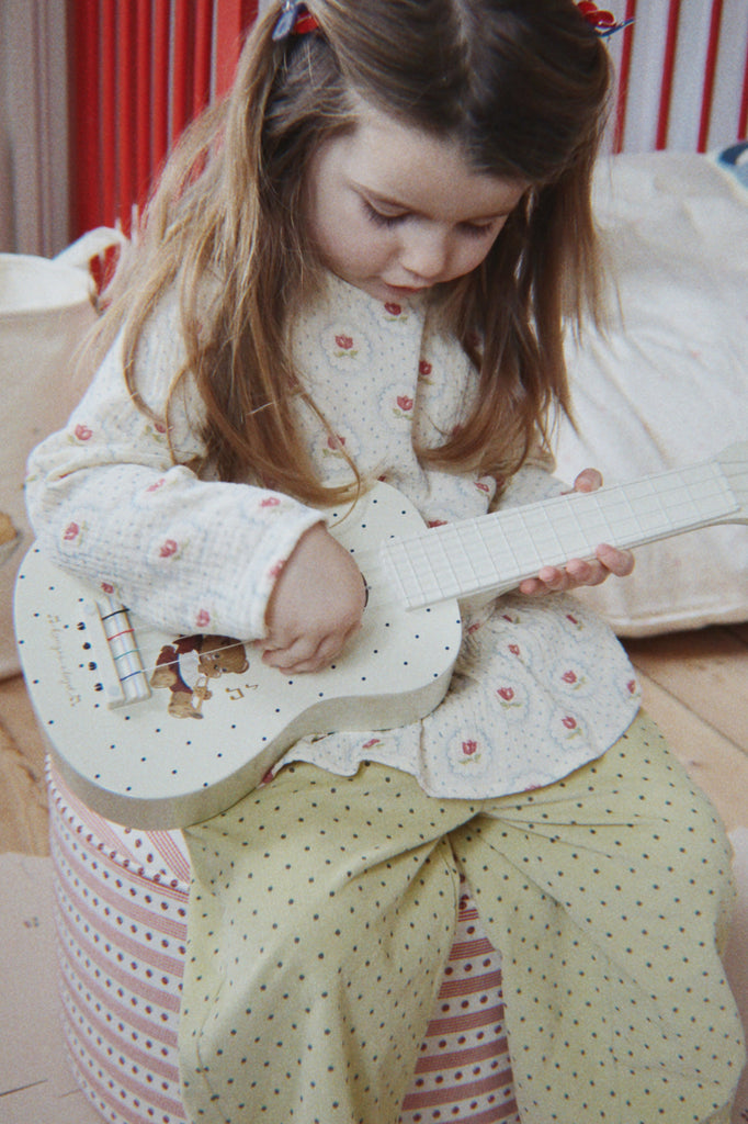 A young girl with long hair sits on a striped pouf and plays the Konges Sloejd Wooden Ukulele - Teddy Bear, a charming musical instrument for kids. She wears a cream top with pink flowers and yellow pants with black polka dots.