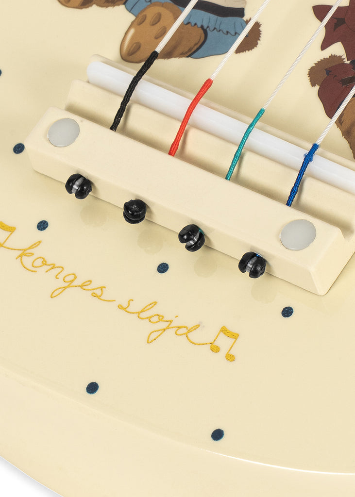 Close-up of the Konges Sloejd Wooden Ukulele - Teddy Bear, showcasing its cream-colored wooden bridge with vibrant colored strings, small black knobs, blue dots, and charming bear illustrations. This delightful kids’ musical instrument features “Konges Sløjd” elegantly written in gold cursive.