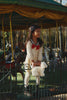 A young girl in a polka-dot dress holds a white stuffed bear and the Konges Sloejd Puppy Bag - Off White while standing on a fence near a carousel, surrounded by trees and sunlight.