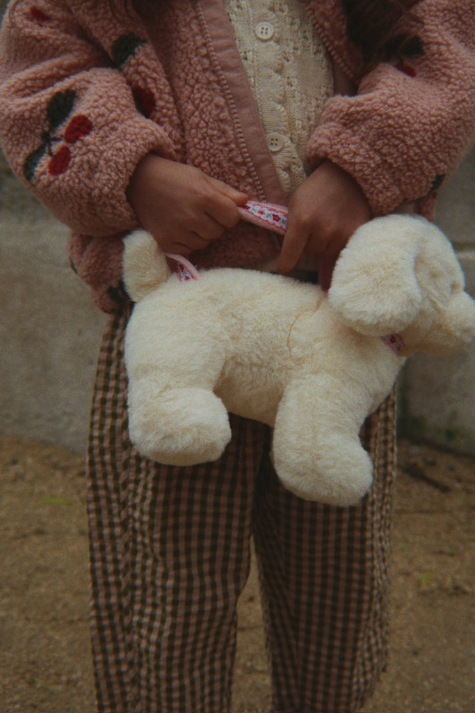 A child dressed in a pink jacket and plaid pants carries the Konges Sloejd Puppy Bag - Off White by its leash.