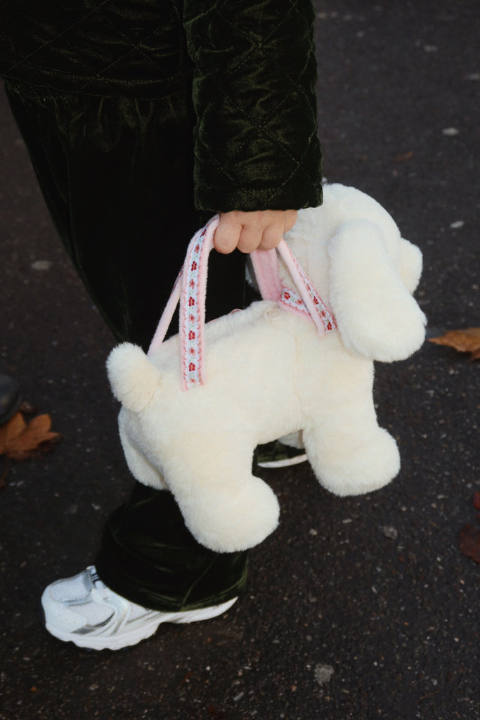 A child in dark clothes and white sneakers holds the Konges Sloejd Puppy Bag - Off White, standing on a leaf-strewn paved surface.