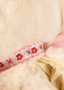 Close-up of the Konges Sloejd Puppy Bag - Off White’s cream fur with a white ribbon adorned with red flowers and green leaves, plus glimpses of light pink fabric and a cream zipper.