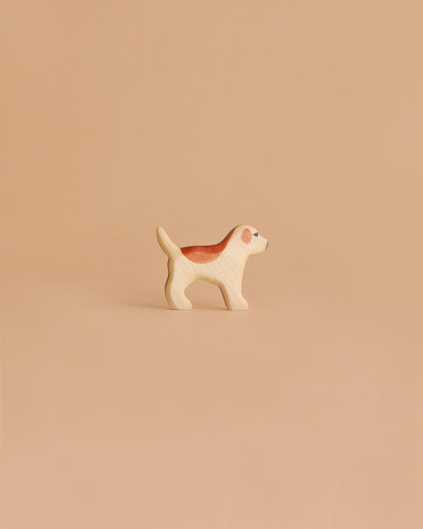 The Handmade Holzwald Golden Retriever Puppy, a small wooden dog figurine with brown and white details, stands on a smooth light brown surface against a beige background, highlighting the charm of sustainable toys.