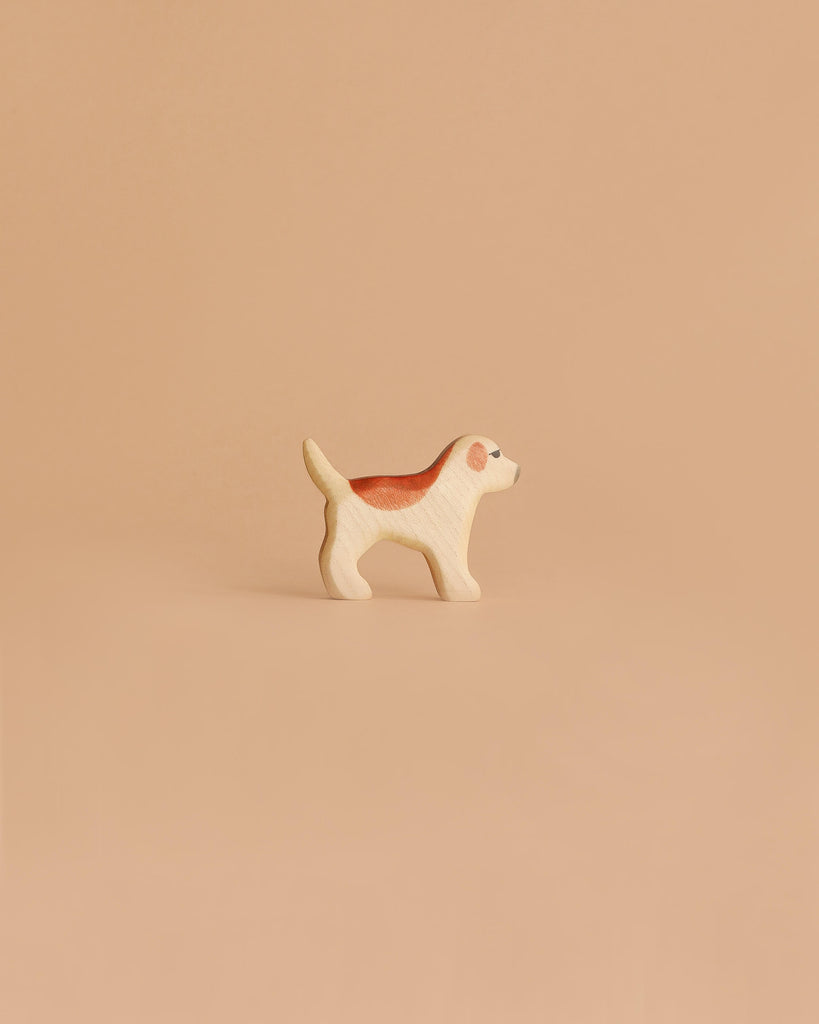 The Handmade Holzwald Golden Retriever Puppy, a small wooden dog figurine with brown and white details, stands on a smooth light brown surface against a beige background, highlighting the charm of sustainable toys.
