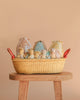 The Maileg Easter Basket Gift Set, featuring fabric bunny dolls and decorative eggs, is displayed in a woven basket on a wooden stool against a beige background.