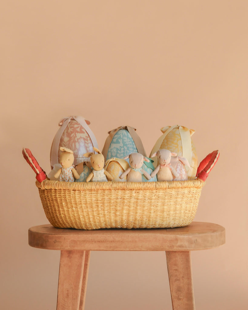 The Maileg Easter Basket Gift Set, featuring fabric bunny dolls and decorative eggs, is displayed in a woven basket on a wooden stool against a beige background.