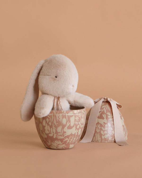 A Maileg Easter Egg with Bunny is displayed in a decorative beige bowl with floral patterns, beside a matching round gift box tied with a beige ribbon, all set against a beige background.