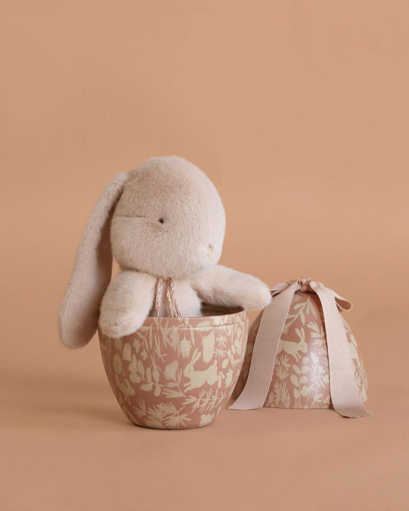 A Maileg Easter Egg with Bunny is displayed in a decorative beige bowl with floral patterns, beside a matching round gift box tied with a beige ribbon, all set against a beige background.