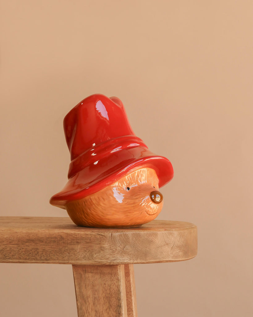 The Paddington Money Bank, a ceramic coin bank shaped like Paddington Bear’s head topped with a red hat, is displayed on a wooden stool against a simple beige background.