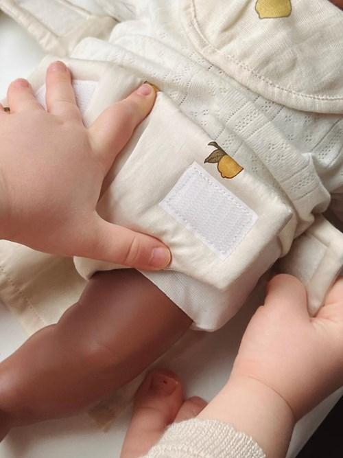 A child fastens the Konges Sloejd Doll Nursery Set - Lemon diaper with velcro onto a brown-skinned baby doll dressed in cream-colored clothes with small yellow lemon designs.