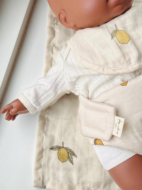 A close-up of a doll on a matching blanket featuring yellow lemons, with its arm outstretched beside the Konges Sloejd Doll Nursery Set – Lemon, which includes organic cotton accessories in coordinating cream and lemon patterns.