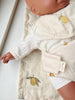 A close-up of a doll on a matching blanket featuring yellow lemons, with its arm outstretched beside the Konges Sloejd Doll Nursery Set – Lemon, which includes organic cotton accessories in coordinating cream and lemon patterns.