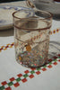 A Konges Sloejd Glitter Cup - Lemon, from the 2 pack set, sits on a tablecloth with colorful checkered embroidery. A plate and another dish are partially visible in the background.