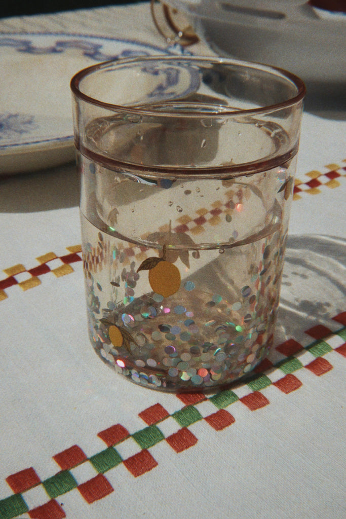 A Konges Sloejd Glitter Cup - Lemon, from the 2 pack set, sits on a tablecloth with colorful checkered embroidery. A plate and another dish are partially visible in the background.