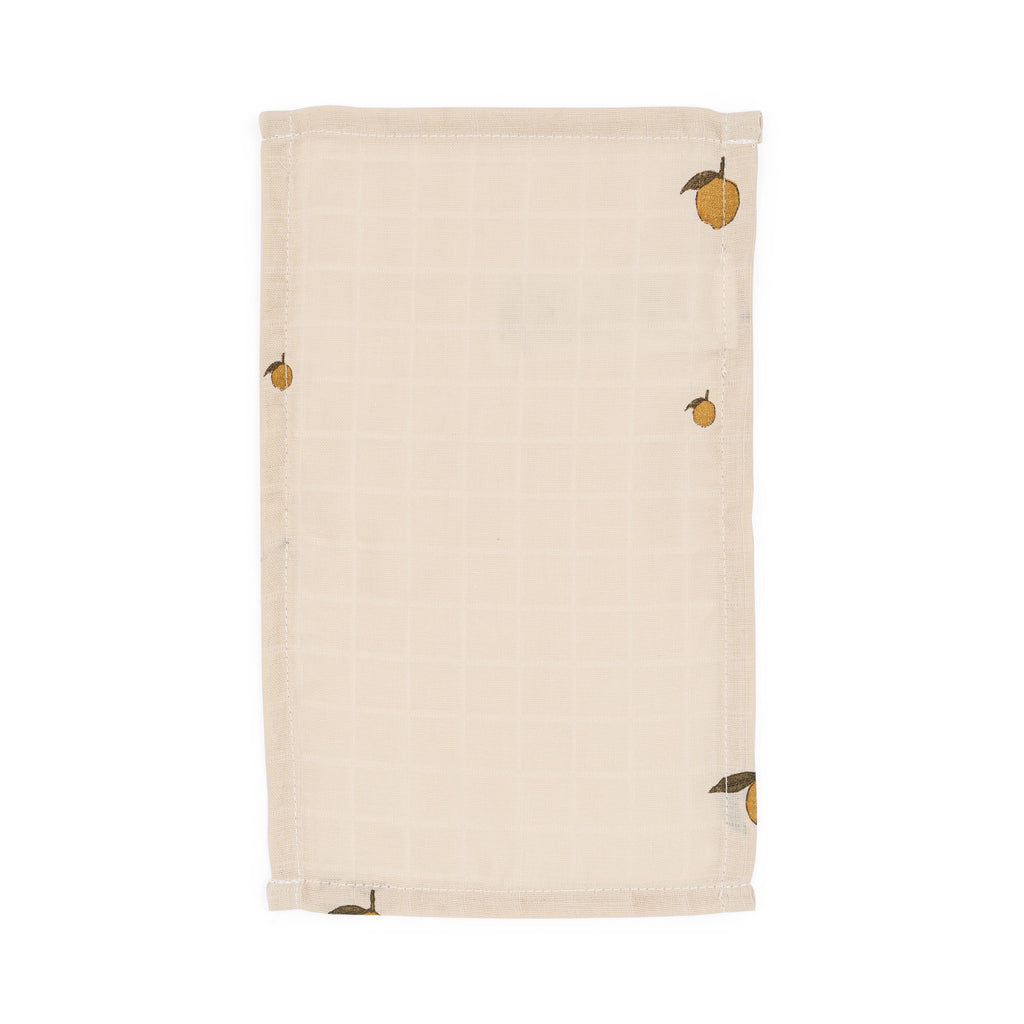 The Konges Sloejd Doll Nursery Set - Lemon features a beige, rectangular organic cotton cloth with a subtle grid and small yellow lemon illustrations with green leaves in three corners, perfect as a doll accessory or bib. Laid flat on white background.