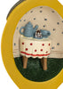 A Konges Sloejd Mini Surprise House - Lemon featuring a miniature world inside a yellow oval frame, with a small table with a polka-dot tablecloth holding a blue teapot, a cup, and a stack of books for imaginative storytelling.